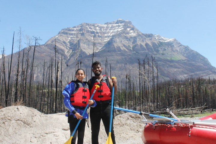 Two people in red vests holding paddles, standing near a raft with mountains in the background.