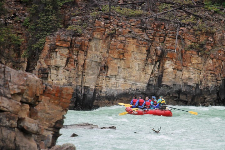 Group in red raft navigating rapids near rocky cliffs.