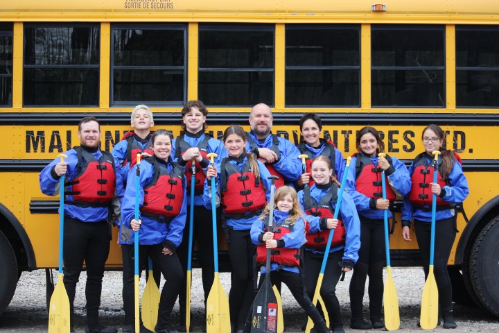 Group of people in red life jackets holding paddles in front of a yellow bus.