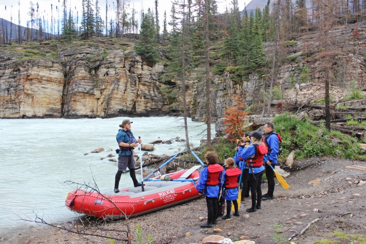 People in life jackets gather near a raft on a rocky riverbank amid tall trees.