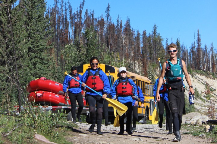 Group in life vests carrying paddles, walking from yellow bus with red rafts in forested area.