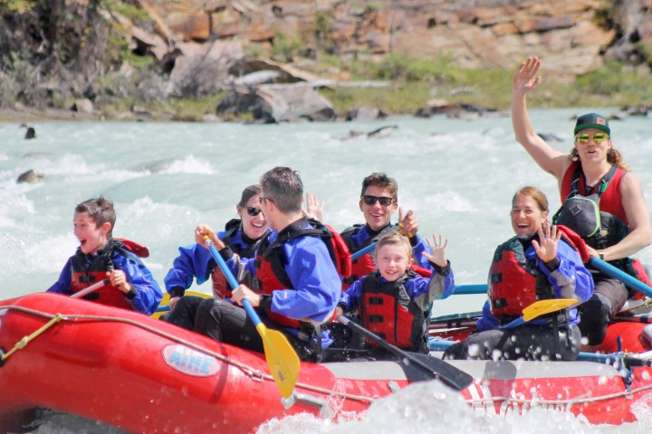 Group rafting on a river, smiling and waving, wearing life jackets and helmets in a red inflatable raft.