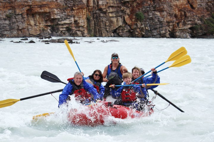 Group of people rafting in whitewater with paddles raised and smiling faces.