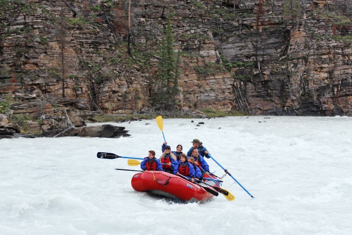 Group whitewater rafting in a red raft on a river with rocky cliff backdrop.