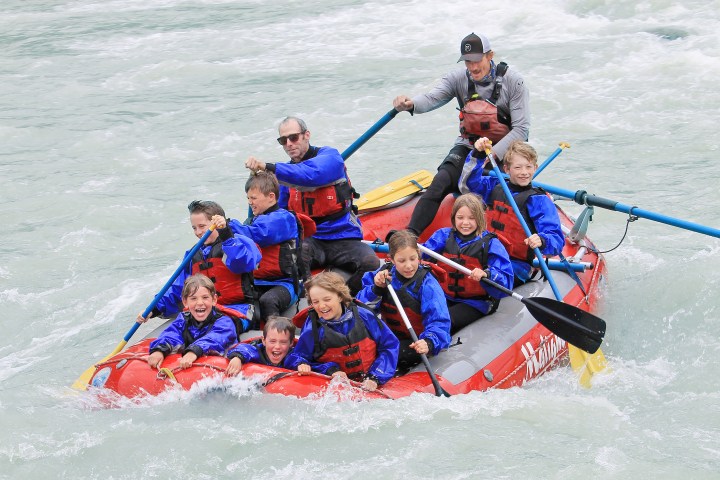 Group of people in blue jackets and helmets enjoying whitewater rafting on a red inflatable raft.