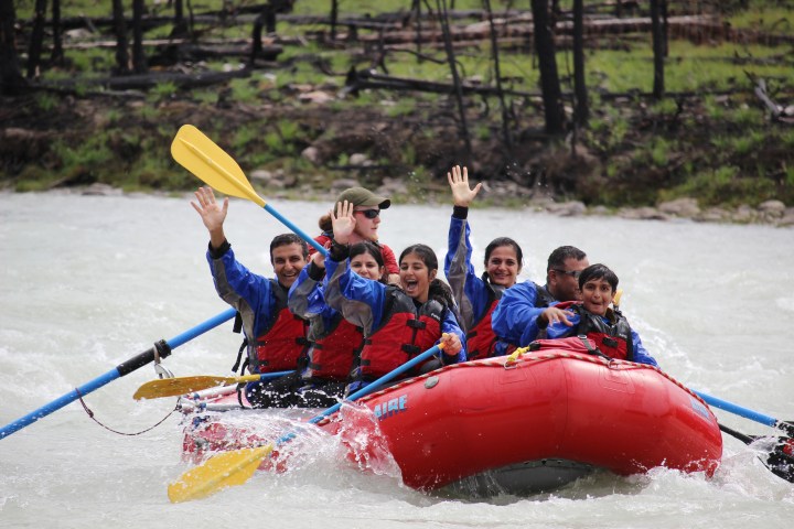 Group of people whitewater rafting in a red inflatable boat, cheering with raised hands.