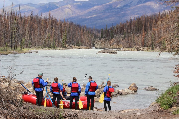 Group preparing for rafting on a river, wearing red life jackets and holding paddles, with trees and mountains in the background.