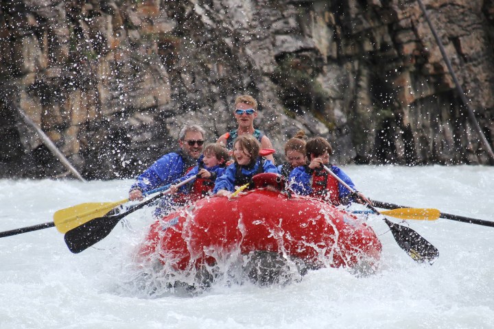 Group of people in a red raft navigating whitewater rapids, splashing water.