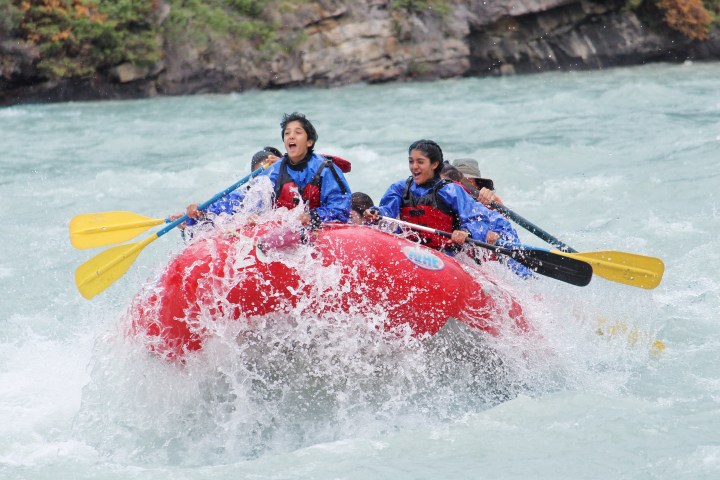 People rafting in a red inflatable boat on a river, splashing through waves.