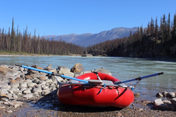 Red inflatable raft on rocky riverbank with mountains and forest in background under clear blue sky.