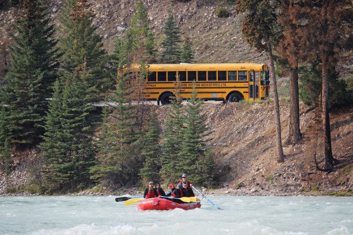 Two people rafting on a river with a yellow school bus in the background.