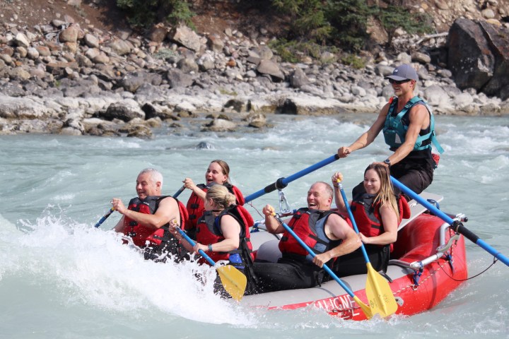 Group of people white-water rafting in a red raft on a fast-moving river.