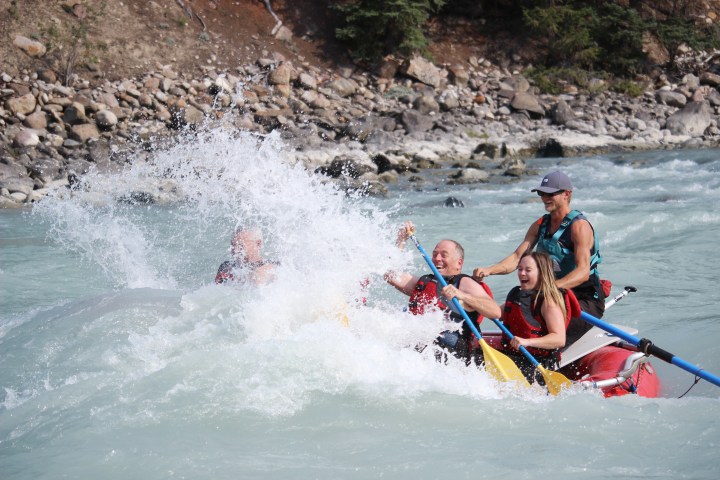 Group rafting on a river, splashing through white water rapids, smiling and holding paddles.