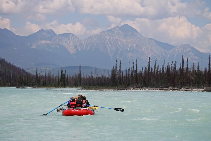 Two people rowing a red raft on a river with mountains in the background.