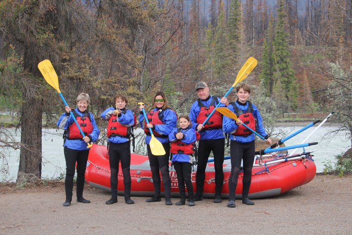 Six people in blue jackets with paddles in front of a red raft beside a river.