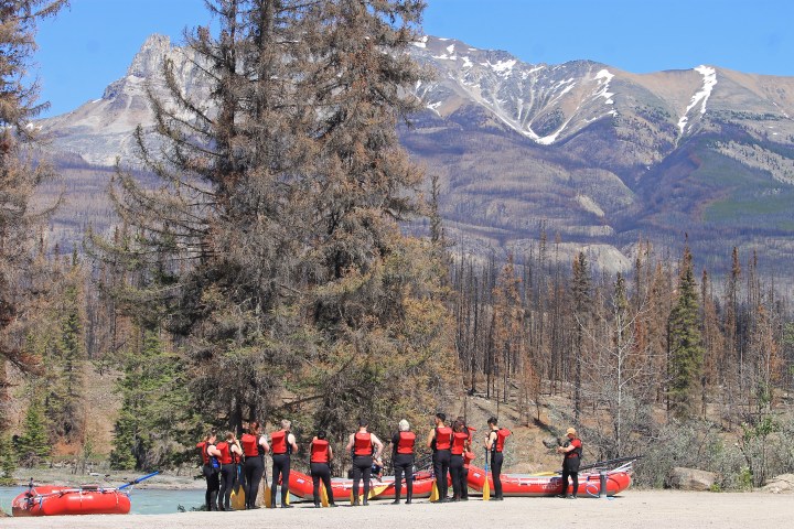 Group of people in red vests with rafts in front of a mountain and forest.