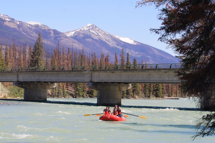 People rafting on a river under a bridge with mountains in the background.
