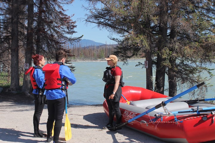 Three people in life jackets beside a red raft near a river, surrounded by trees.